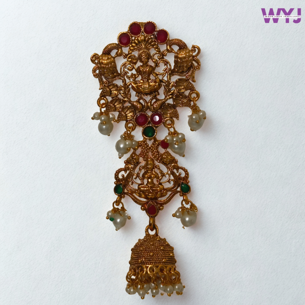 Temple Design Hair Brooch featuring a divine Goddess Lakshmi motif seated in a lotus, flanked by intricate peacocks and floral detailing. Meticulously crafted in antique gold-tone finish, this hair ornament is adorned with red and green stone accents and finished with hanging faux pearl clusters and a jhumka drop for added elegance. Ideal for pairing with sarees or lehengas, especially for weddings, classical dances, and religious ceremonies. This hair brooch is a timeless piece for those who admire rich South Indian temple jewellery aesthetics.